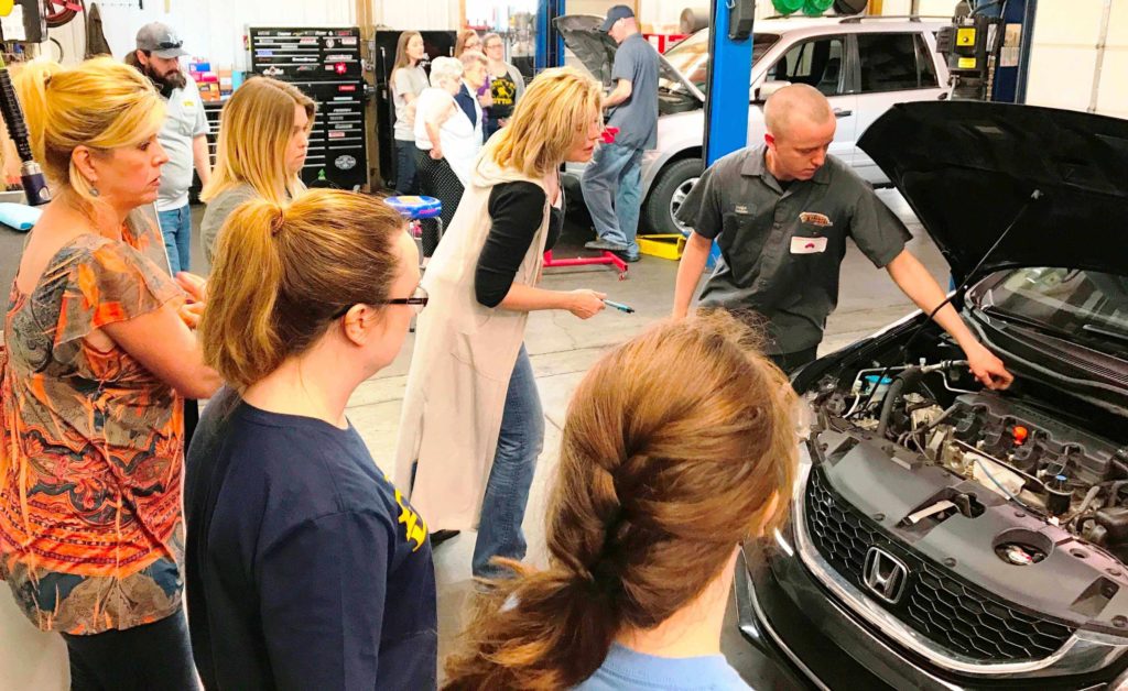Image of a group of women standing around a car that a mechanic is giving them a demonstration on where to find the oil stick to check oil at the American Import Auto Repair Womens Car Care Clinis