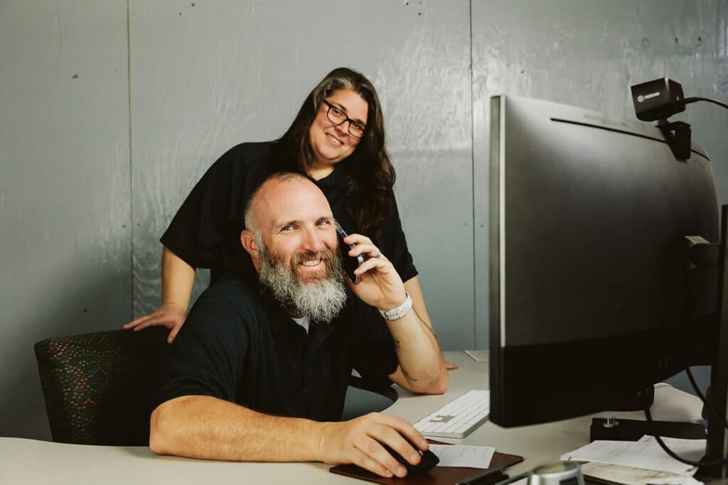Making Great First Impressions With Your Auto Repair Shop with Shop Marketing Pros. Two people in an office, with a man seated at a desk using a phone and a woman standing behind him.